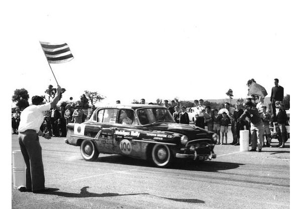 Ein Oldtimer mit der Nummer „100“ auf der Seite fährt bei einer Rennveranstaltung an einem fahnenschwingenden Offiziellen vorbei, der von einer Menschenmenge umgeben ist.