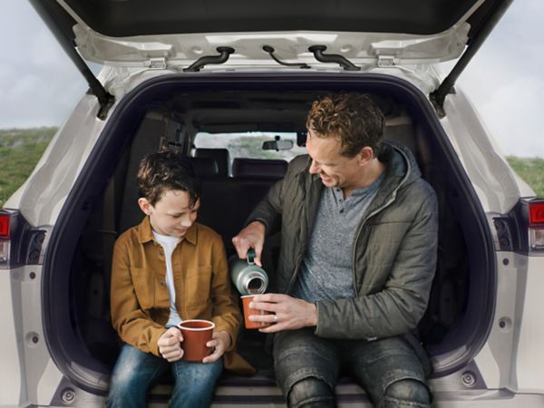 Ein Mann schüttet seinem Sohn, der neben ihm im geöffneten Kofferraum sitzt, ein heißes Getränk aus einer Thermosflasche in den Becher