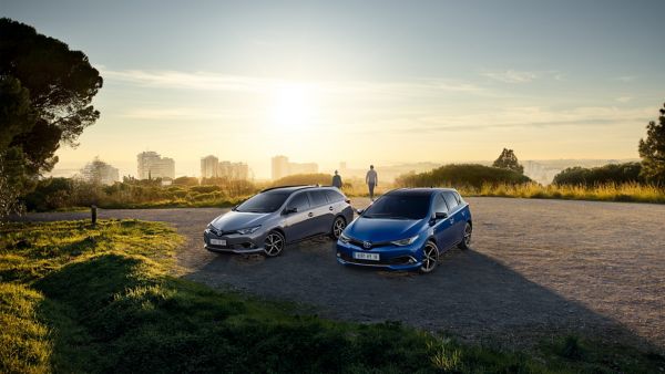 Zwei Autos, ein silbernes und ein blaues, parken bei Sonnenuntergang an einem Aussichtspunkt. Zwei Menschen stehen im Hintergrund und genießen die Aussicht.