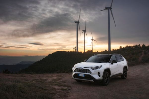 Weißer Toyota RAV4 steht bei Sonnenuntergang auf einer Schotterstraße vor Windkraftanlagen 