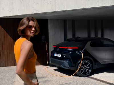Frau mit Sonnenbrille steht vor einem schwarzen C-HR, der unter einer überdachten Einfahrt an einer Wallbox lädt.