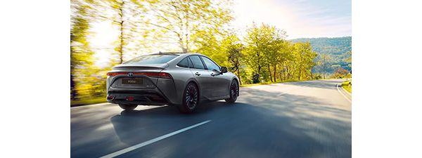 Ein grauer Toyota Mirai fährt auf einer von Bäumen gesäumten Straße. Sonnenlicht scheint durch die Bäume im Hintergrund.