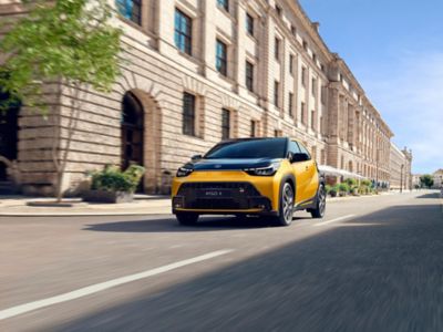 Gelb-schwarzer Aygo X fährt dynamisch durch eine Stadtstraße mit historischen Gebäuden; Bewegungsunschärfe betont die Fahrt.