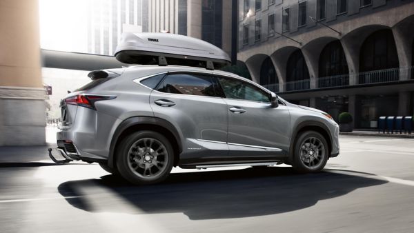 Ein eleganter silberner Lexus mit einer Dachbox, fährt auf einer städtischen Straße, umgeben von moderner Architektur und natürlichem Licht.