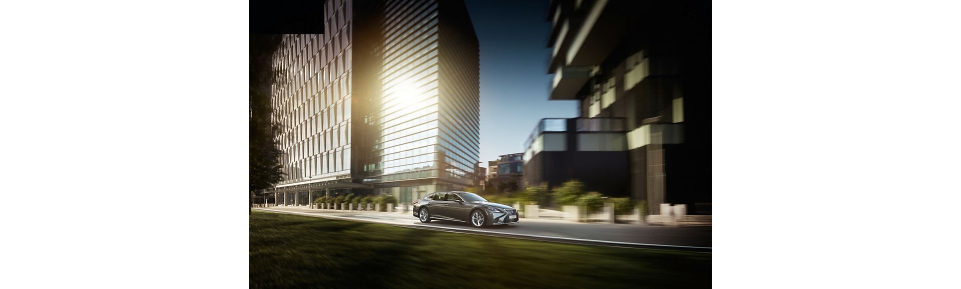 Eine silberne Lexus-Limousine fährt eine malerische Stadtstraße entlang, flankiert von modernen Gebäuden und Grünanlagen