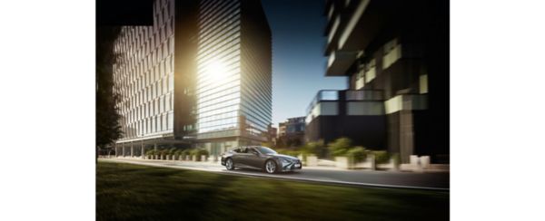Eine silberne Lexus-Limousine fährt eine malerische Stadtstraße entlang, flankiert von modernen Gebäuden und Grünanlagen