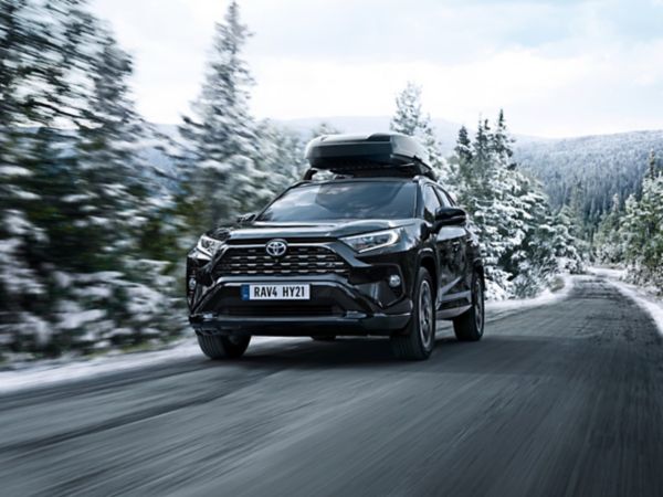 Dunkelgrauer RAV4 mit Dachbox schräg von vorne dynamisch fotografiert, auf einer Straße durch einen winterlichen Nadelwald fahrend 