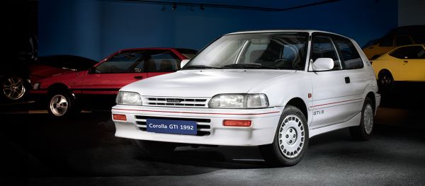 Weißer Corolla GTi von 1992 mit rotem Dekorstreifen schräg von vorn im Spot-Licht in einer abgedunkelten Ausstellungshalle, im Hintergrund weitere Modelle