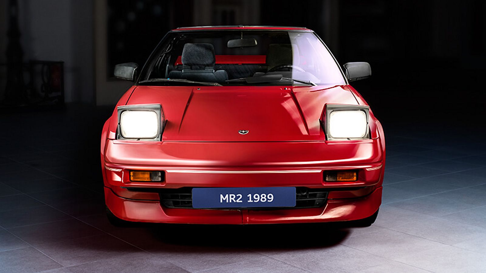 Frontansicht eines roten Toyota MR2 von 1989 mit hochgeklappten Scheinwerfern, punktuell von oben beleuchtet vor dunklem Hintergrund 