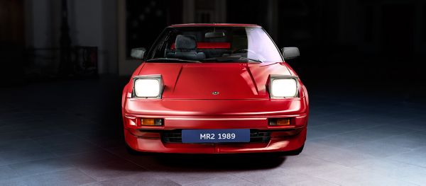 Frontansicht eines roten Toyota MR2 von 1989 mit hochgeklappten Scheinwerfern, punktuell von oben beleuchtet vor dunklem Hintergrund 