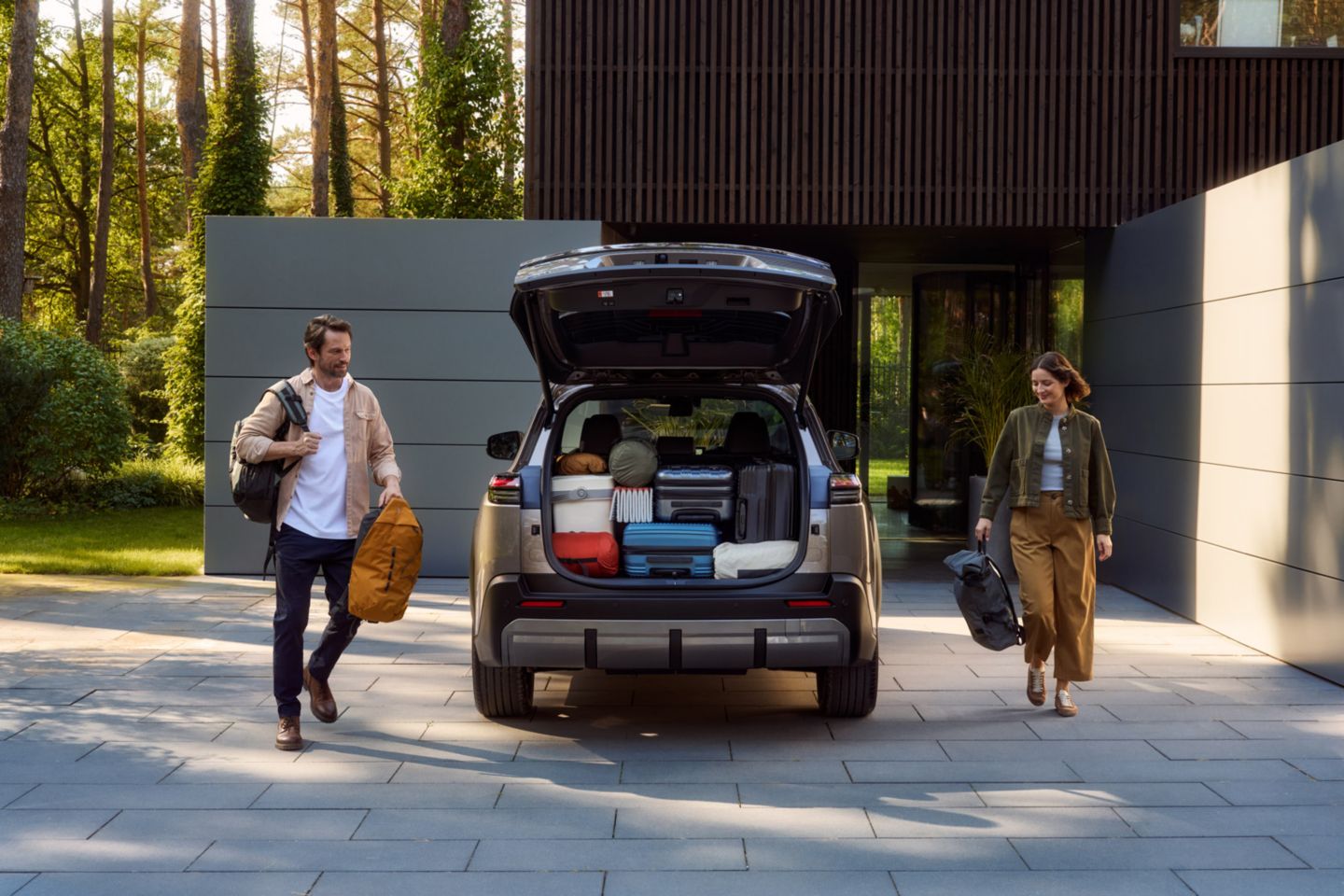 Zwei Personen gehen von einem Haus zu einem geparkten Auto mit geöffneter Heckklappe, das mit Koffern, Taschen und Decken voll beladen ist; beide tragen weitere Gepäckstücke in den Händen.