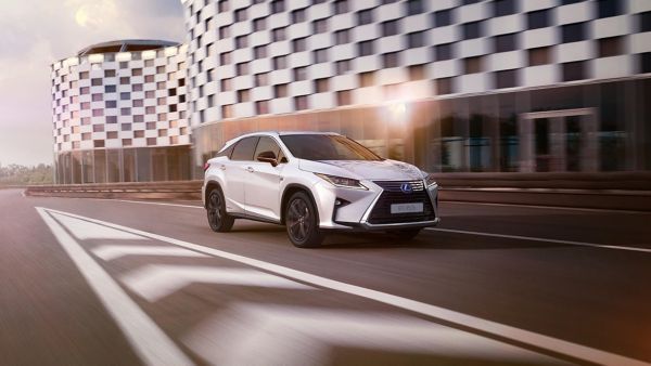Ein eleganter weißer Lexus fährt auf einer Straße neben modernen, gemusterten Gebäuden. Sanftes Abendlicht unterstreicht die dynamische Bewegung des Fahrzeugs.