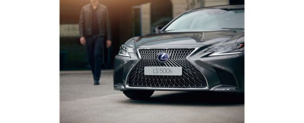 Ein eleganter grauer Lexus LS ist gut sichtbar mit seinem markanten Kühlergrill abgebildet. Eine Person in einer dunklen Jacke nähert sich dem Auto in einer städtischen Umgebung.