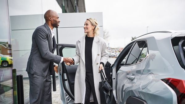 Ein Mann im Anzug schüttelt einer Frau im Mantel die Hand, während sie lächelnd aus einem Lexus steigt.