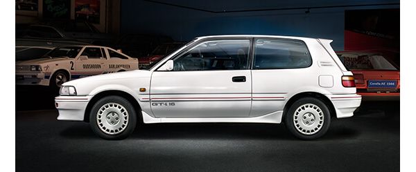 Seitenansicht eines weißen Corolla GTi von 1992 mit rotem Dekorstreifen und GTi-Sticker in einer Ausstellungshalle, im Hintergrund weitere Modelle