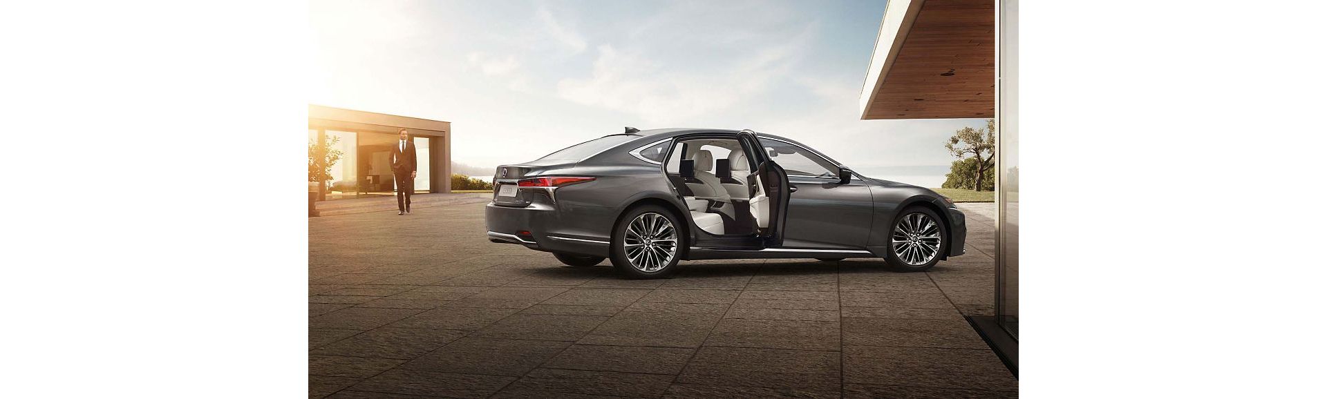 Ein grauer Lexus LS parkt neben einem modernen Gebäude, die hintere Tür ist geöffnet und gibt den Blick auf einen geräumigen Innenraum frei. Ein Mann in einem Anzug nähert sich dem Auto.