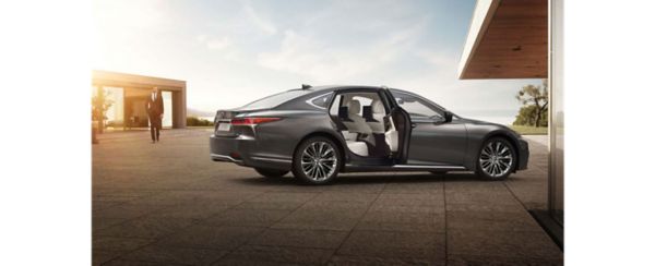Ein grauer Lexus LS parkt neben einem modernen Gebäude, die hintere Tür ist geöffnet und gibt den Blick auf einen geräumigen Innenraum frei. Ein Mann in einem Anzug nähert sich dem Auto.