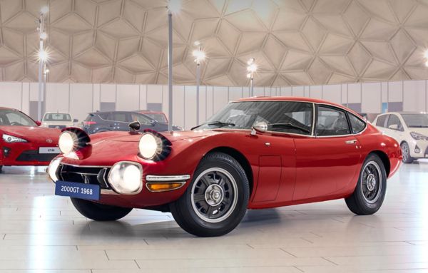 Ein roter Toyota 2000GT von 1968 mit leuchtenden Scheinwerfern in einer hell ausgeleuchteten Ausstellungshalle, im Hintergrund weitere Modelle
