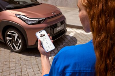 Person hält ein Smartphone mit der geöffneten MyToyota App in der Hand; im Hintergrund die Front eines Fahrzeugs.