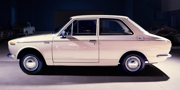 Seitenansicht eines crèmefarbenen Corolla von 1967 in einer gefliesten Ausstellungshalle, punktuelles Licht, dunkler Hintergrund
