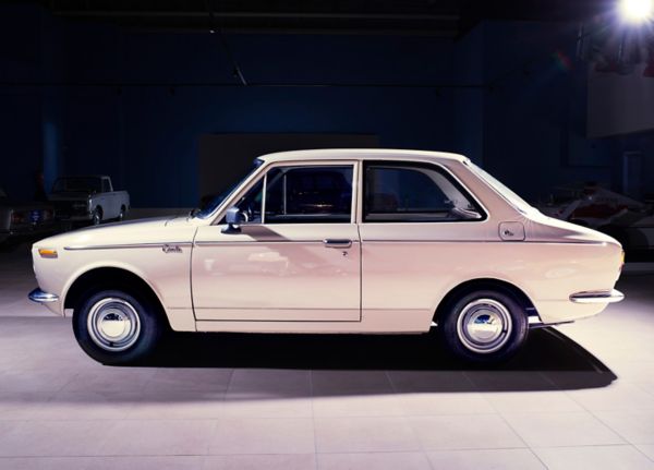 Seitenansicht eines crèmefarbenen Corolla von 1967 in einer gefliesten Ausstellungshalle, punktuelles Licht, dunkler Hintergrund