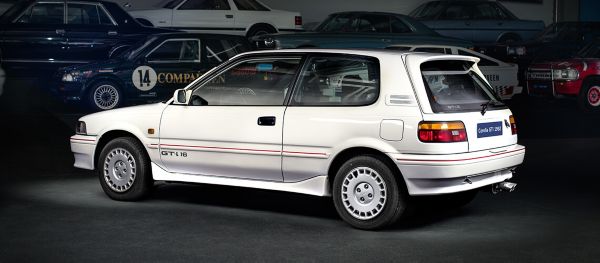 Weißer Toyota Corolla GTi aus dem Jahr 1992 schräg von hinten im Spot-Licht in einer abgedunkelten Ausstellungshalle, im Hintergrund weitere Fahrzeuge