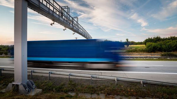 Auf dem Bild ist ein schnell fahrender blauer Lkw zu sehen, der auf einer Autobahn unter einem Maut- oder Überwachungssystem erfasst wird.