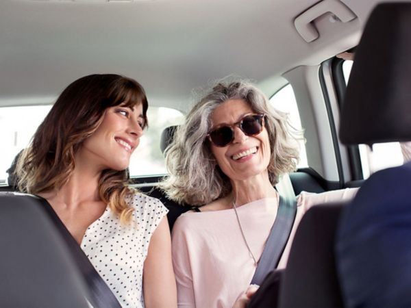 Zwei Frauen in einem Auto teilen einen freudigen Moment, sie lächeln sich an, während sie angeschnallt sind. Eine Frau hat langes Haar und die andere hat lockiges graues Haar.