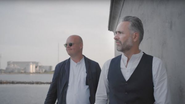 Zwei Männer mit Hemd und Weste bzw. Sakko blicken vor einer grauen Betonwand aufs Meer