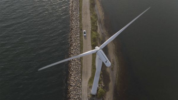 Luftaufnahme eines Windrads auf einem schmalen Landstreifen, rechts und links Wasser, davor geparkt ein weißes Fahrzeug