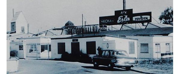 Schwarz-Weiß-Foto aus den 60er-Jahren: ein flaches Industriegebäude, auf dem Dach ein Schild, unter anderem mit den Schriftzügen „Erla“ und „Toyota“