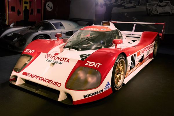 Weiß-roter TS010 Rennwagen mit Toyota Schriftzug und verschiedenen Sponsoren-Logos von schräg vorn, angestrahlt in einem dunklen Ausstellungsraum