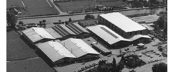 Schwarz-Weiß-Foto aus den 60er-Jahren: Luftaufnahme eines Industriegeländes mit mehreren Hallen und einem Parkplatz nahe eines Flusses