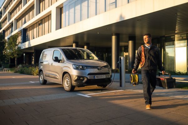 Ein Toyota Proace Electric an der Ladestation. Davor sein Besitzer mit Werkzeugkasten in der linken Hand und Ladekabel um seine Schulter.