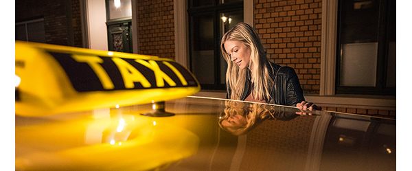 Im Vordergrund das Dach eines Taxis mit einem leuchtenden Taxi-Schild. Eine Frau steigt in das Taxi ein.