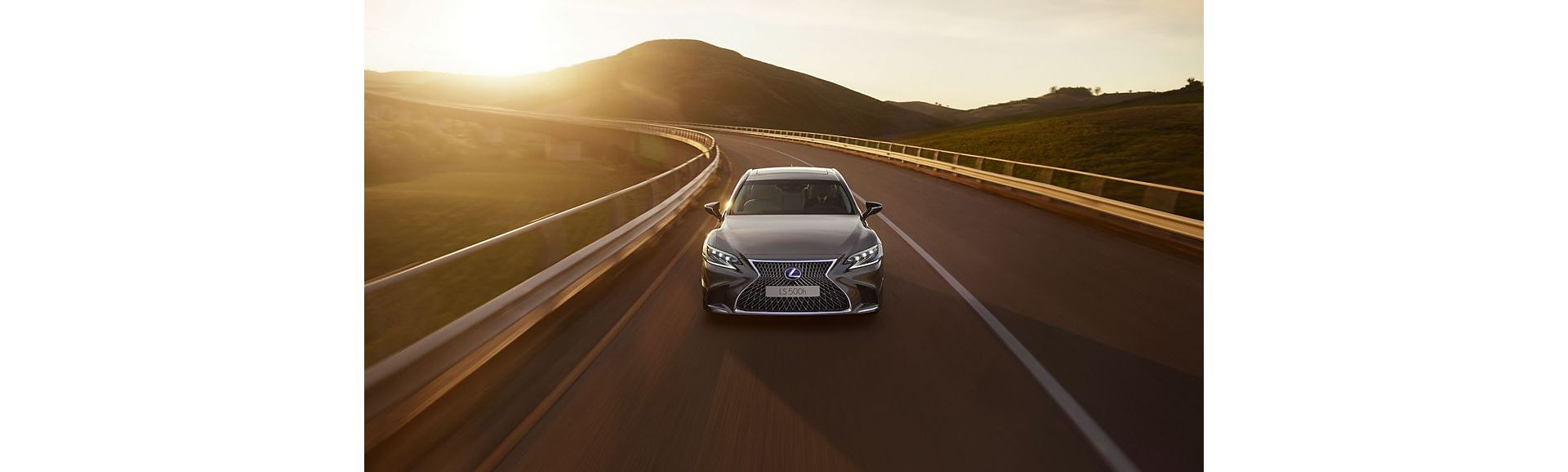 Ein eleganter grauer Lexus LS auf einer kurvenreichen Straße, umgeben von sanften Hügeln bei Sonnenuntergang
