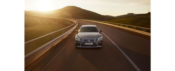 Ein eleganter silberner Lexus fährt auf einer kurvenreichen Straße, umgeben von üppig grünen Hügeln, die von einem warmen Sonnenuntergang beleuchtet werden.