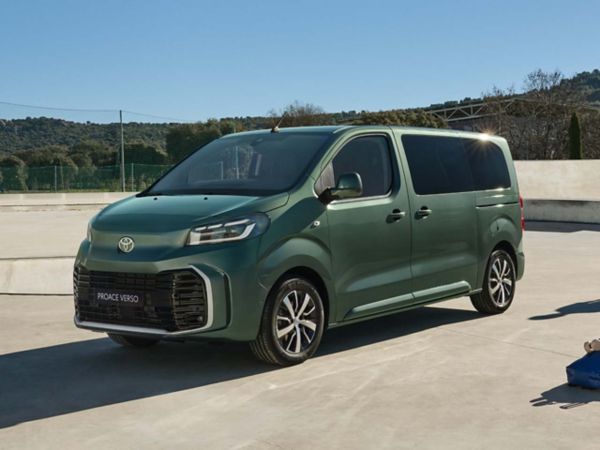 Ein grüner Toyota Proace Verso, der an einem sonnigen Tag auf einer flachen Betonfläche parkt mit Bäumen umringt. 