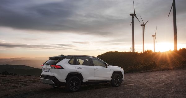 Ein weißer Toyota RAV4 SUV bei Sonnenuntergang auf einem rauen Terrain, im Hintergrund sieht man einen Windpark