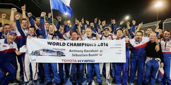 Eine Gruppe jubelnder Mitglieder des Toyota-Rennteams feiert ihren Sieg bei der Weltmeisterschaft 2014 und hält ein Banner mit der Aufschrift „WORLD CHAMPIONS 2014“ und den Namen der Rennfahrer.