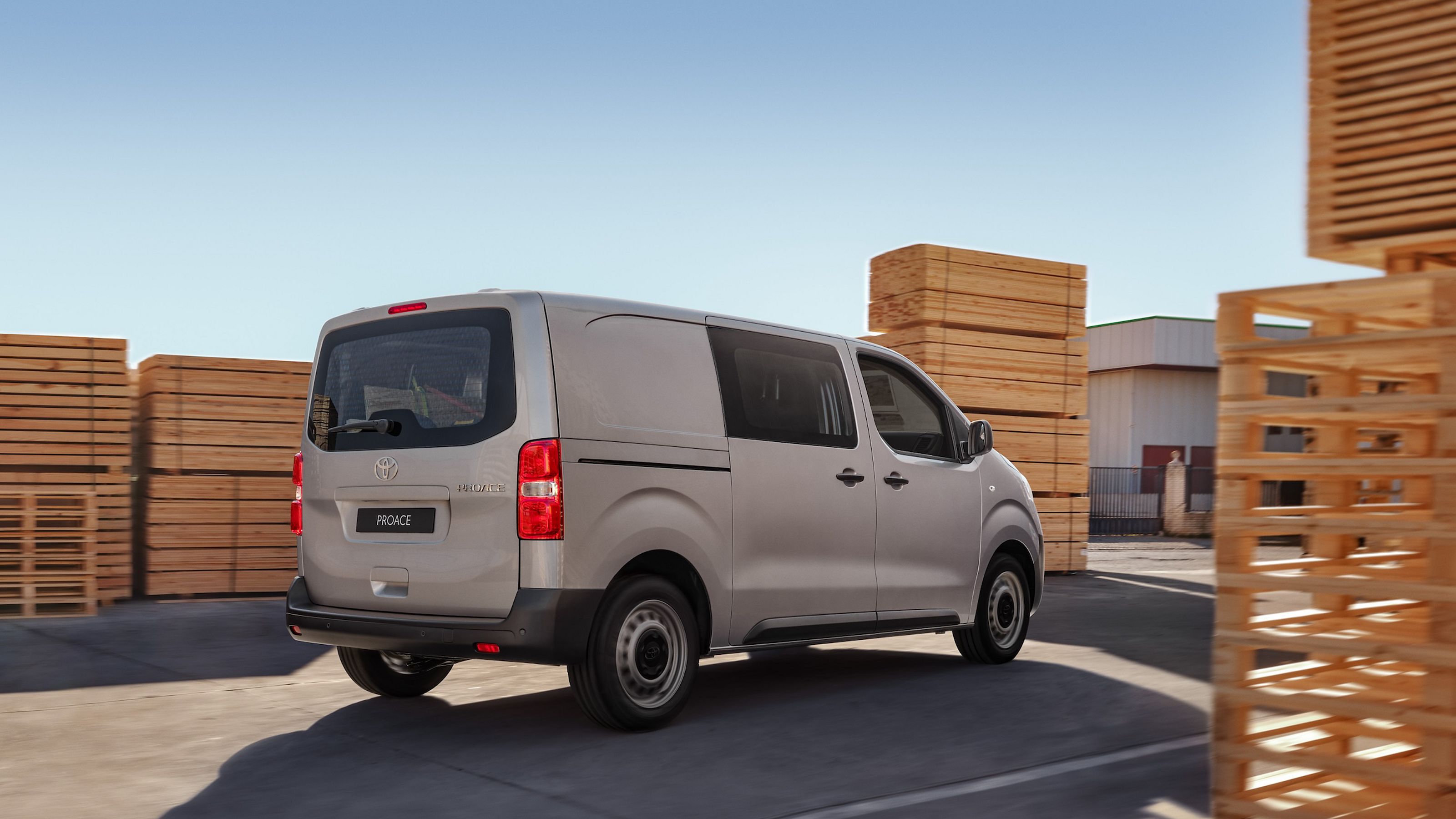 Ein grauer Proace-Transporter schräg von hinten auf einem Industriegelände fahrend, links und rechts Stapel mit Paletten und anderen Holzteilen