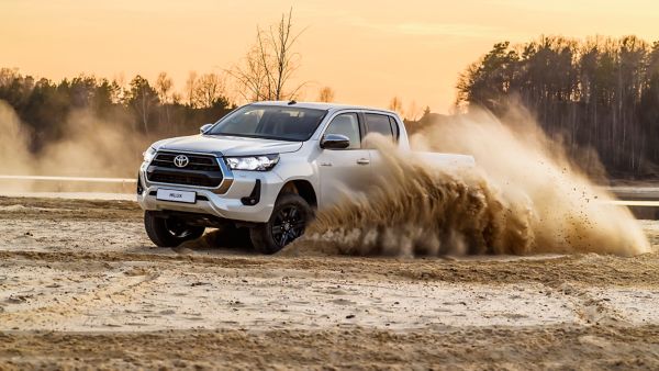 Ein weißer Hilux driftet auf einer Sandfläche und wirbelt eine große Menge Staub und Sand auf, im Hintergrund ein Wald