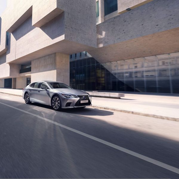 Eine elegante silberne Lexus LS Limousine fährt auf einer modernen Stadtstraße mit dramatischen architektonischen Strukturen im Hintergrund.