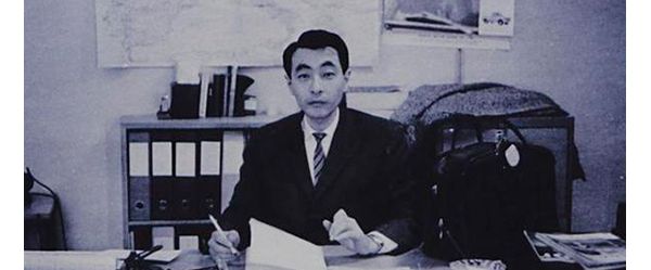 Schwarz-Weiß-Foto: Ein japanischer Mann mittleren Alters sitzt mit Anzug und Krawatte an einem Schreibtisch und blickt in die Kamera