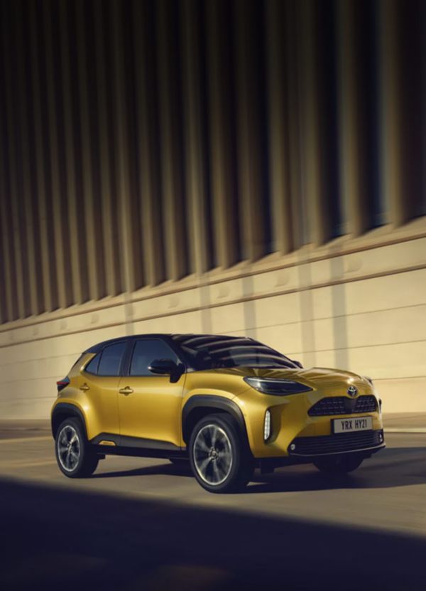 Frontale Seitenansicht eines gelben Toyota Yaris Cross Hybrid, der an einer unscharfen Gebäudefassade entlang fährt.