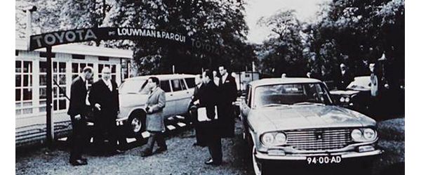 Schwarz-Weiß-Foto aus den 60er-Jahren: Eine Gruppe von Männern zwischen geparkten Autos vor einem Schild mit dem Schriftzug „Toyota Louwman & Paraqui“