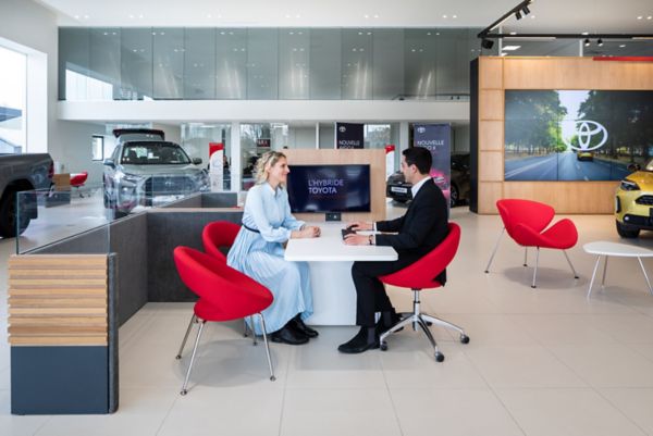 Ein Mann in einem Anzug und eine Frau in einem blauen Kleid sitzen an einem Tisch in einem modernen Autohaus und besprechen Fahrzeugoptionen. Rote Stühle und Autos im Hintergrund unterstreichen die Szenerie.