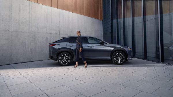 Eine Person in elegantem Schwarz gekleidet geht an einem dunkelgrauen Lexus vorbei, der vor einem Haus geparkt ist