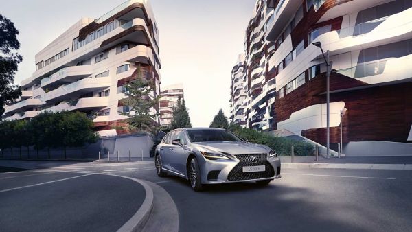 Ein silberner Lexus LS fährt auf einer kurvigen Straße, die von modernen Wohnhäusern mit einzigartigem architektonischem Design umgeben ist.