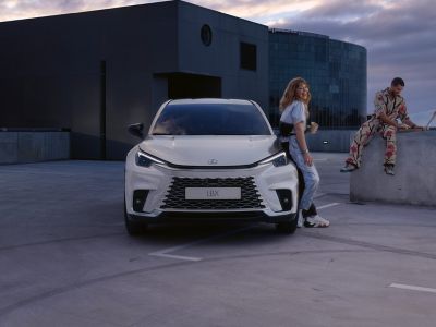 Ein eleganter weißer Lexus LBX, der auf einem Dach geparkt ist, mit einer Frau, die lässig daneben steht, und einem Mann, der auf einer Betonbarriere sitzt